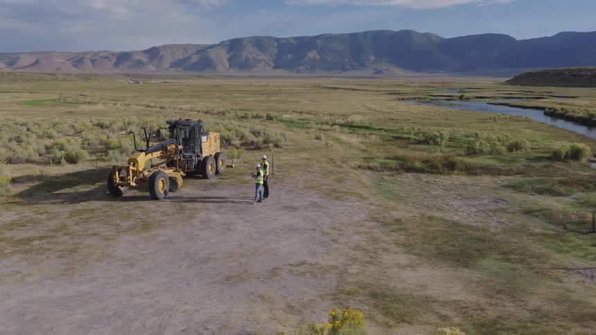 Aerial shot of two construction workers surveying a remote site next to a road grader in a picturesque mountain valley