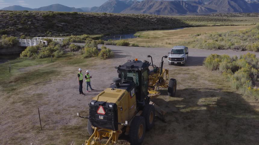 High angle crane shot of two construction workers standing by a road grader and discussing plans on a rural job site with mountains in the distance