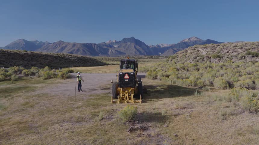 An aerial drone circles two construction workers as they survey a rural job site with a road grader and a vast mountain range in the background