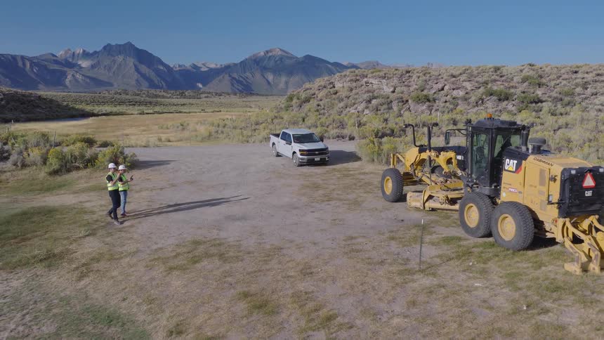 Aerial view pulling away from two engineers surveying a rural construction site with a road grader and pickup truck under a vast mountain range