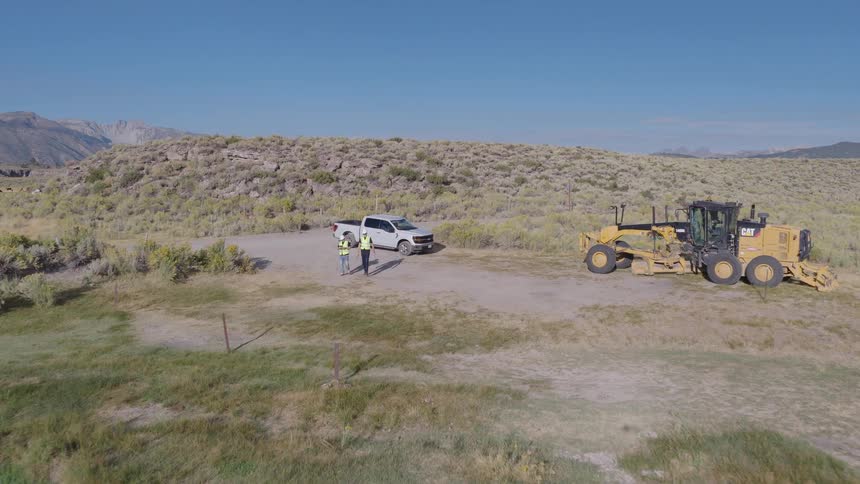 Aerial view of two surveyors in hard hats discussing plans at a rural worksite next to a river and heavy construction equipment