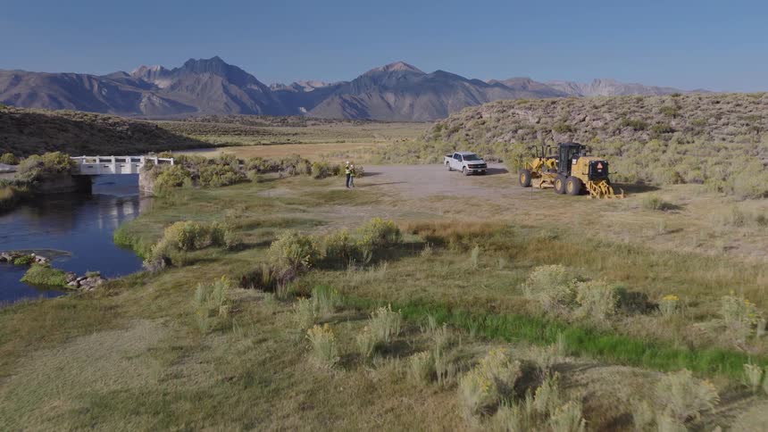 Aerial view slowly pushes in on two surveyors at a rural construction site with a grader and truck by a river, with majestic mountains in the distance