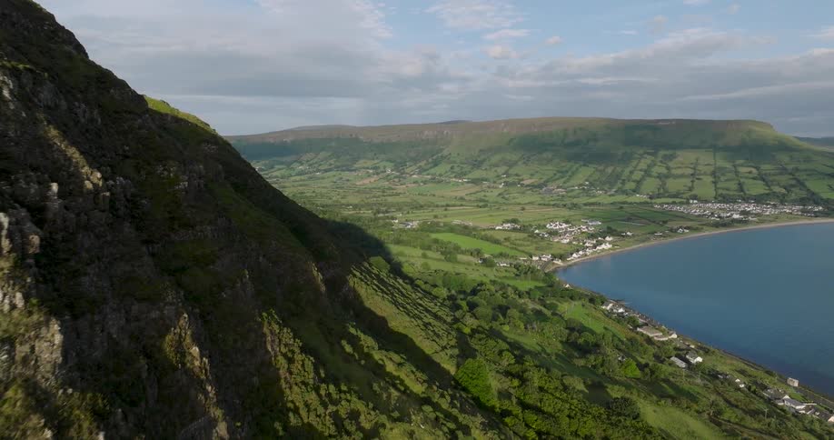 An epic aerial dolly shot reveals the stunning green coastal landscape of Northern Ireland, with dramatic cliffs overlooking a serene bay and a quaint seaside village