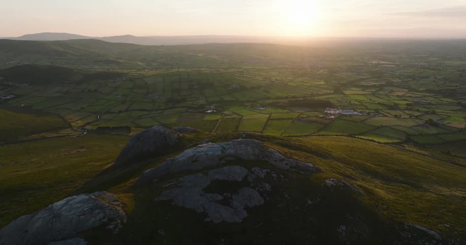Majestic aerial view from a rocky mountain peak overlooking a sprawling valley of green, patchwork fields as the sun sets on the horizon