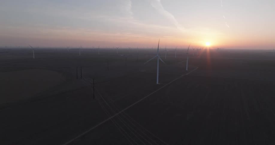 A slow aerial pullback reveals an expansive wind farm with dozens of turbines spinning against a beautiful sunrise