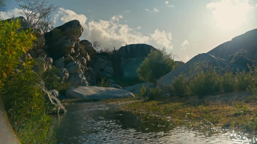 A tranquil stream gently flows through a sunlit rocky landscape under a partly cloudy sky