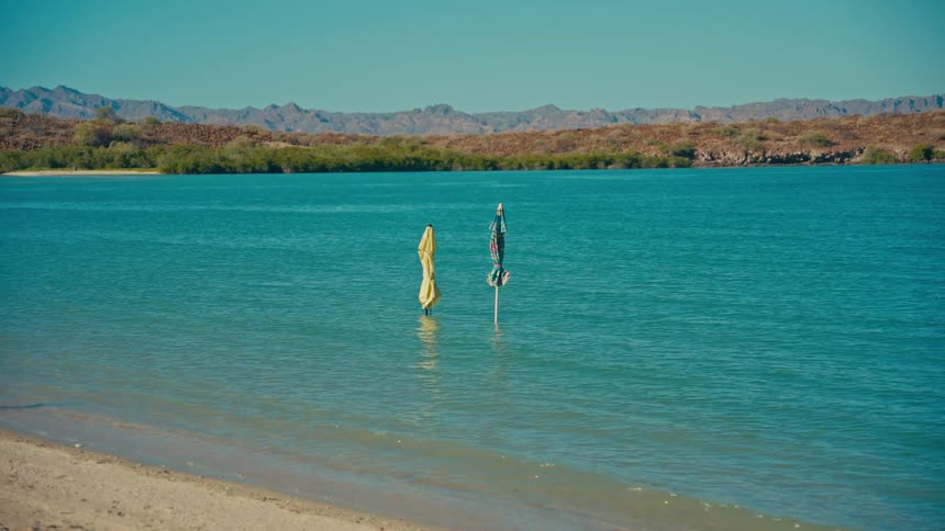 Two closed beach umbrellas stand in the calm turquoise water of a tranquil bay on a sunny day