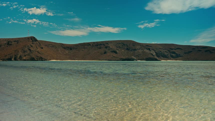 A serene, static view of crystal-clear shallow ocean water with a dry, mountainous island in the background under a blue sky