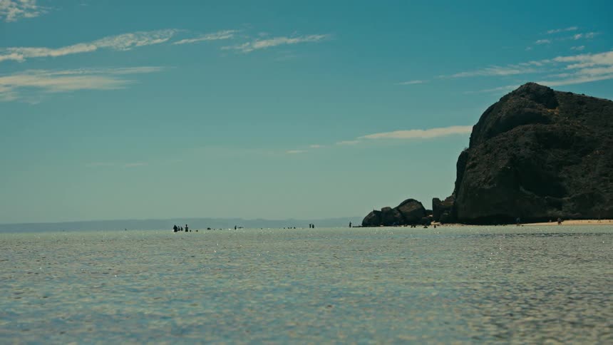 A beautiful, static shot of a calm, clear ocean with a rocky cliff and distant people on a sunny beach