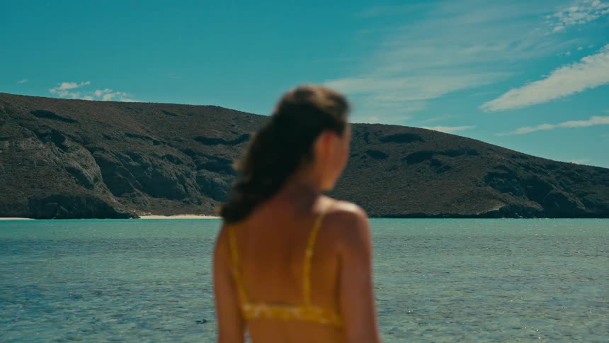 A beautiful tropical landscape with a calm turquoise ocean and rocky hills is seen beyond an out-of-focus woman in a yellow bikini