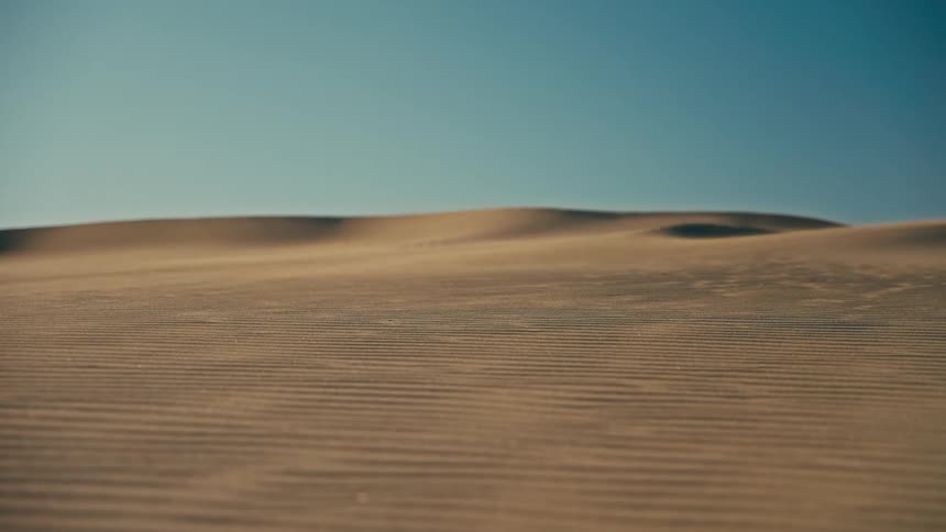 A desert wind blows fine sand over rippled dunes under a clear blue sky