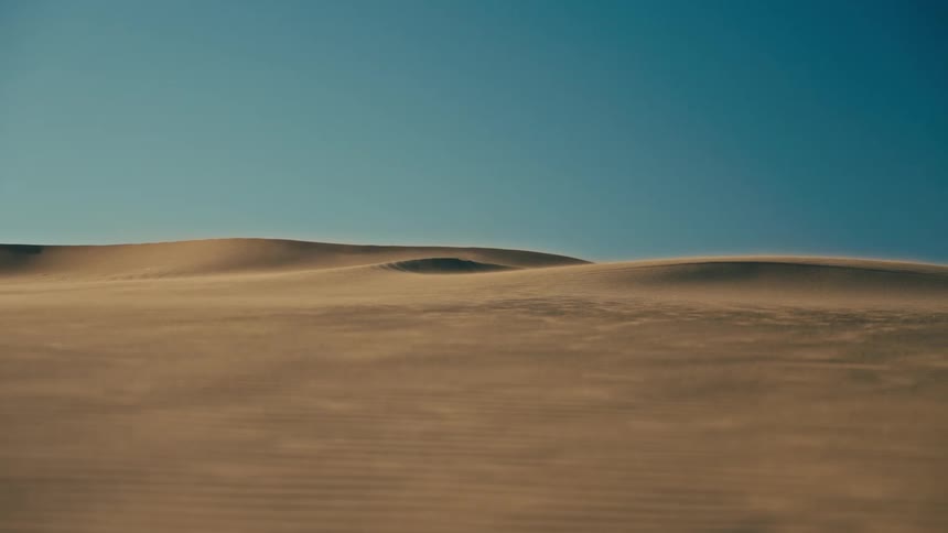 Wind sweeps across vast desert sand dunes under a clear, deep blue sky