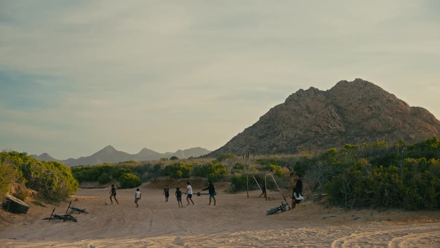 A group of kids play a slow-motion game of soccer on a makeshift field in a sandy, mountainous landscape