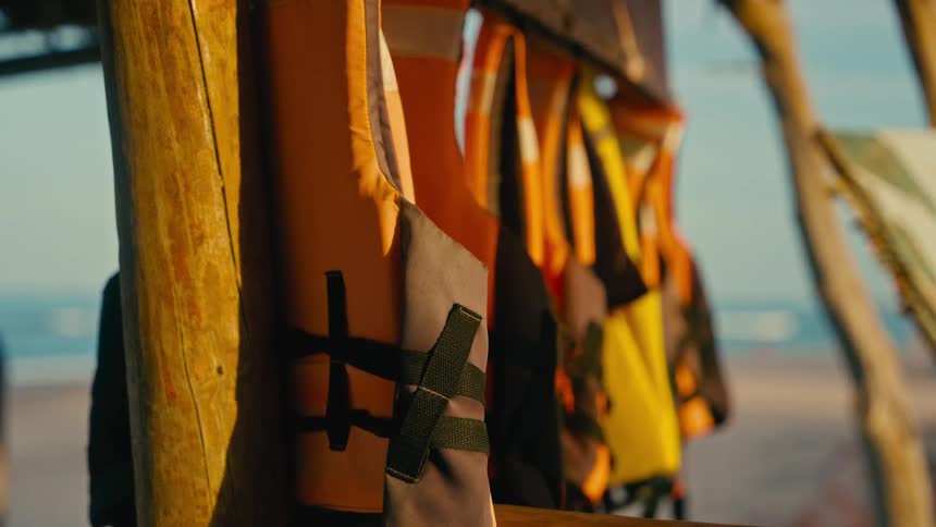 A rack of colorful life jackets hangs in a rustic beach hut during a warm, golden sunset with the ocean in the background