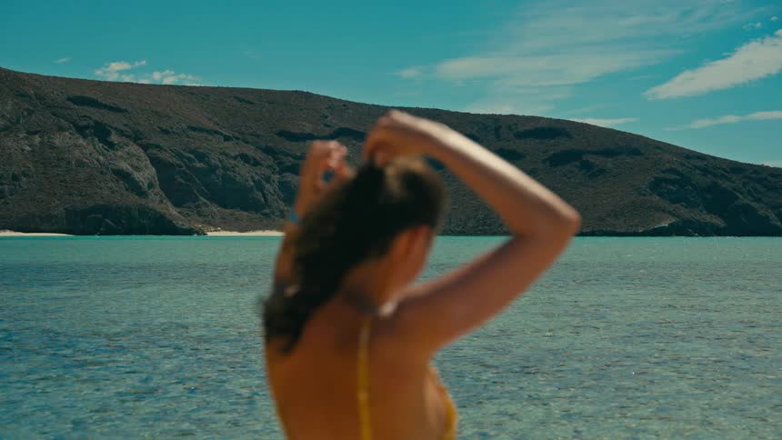 A woman in a yellow bikini ties her hair back in slow motion, with the stunning turquoise waters of a secluded bay in the background