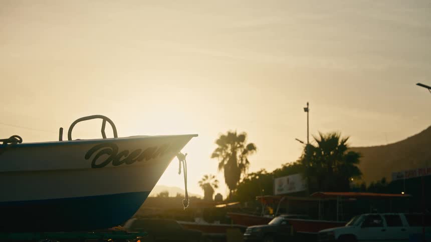 The bow of a small fishing boat named 'Ocean' sits on land, silhouetted against a warm, golden sunset in a coastal village