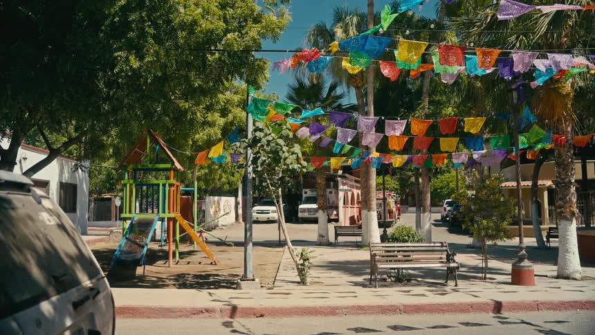 Colorful papel picado banners hang over a quiet street with a children's playground in a sunny small town