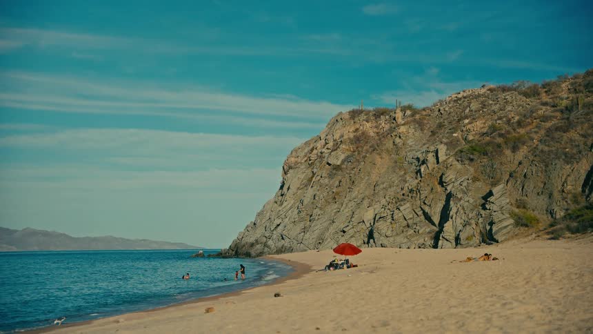 A serene wide shot of people enjoying a sunny day on a sandy beach nestled against a rocky cliff