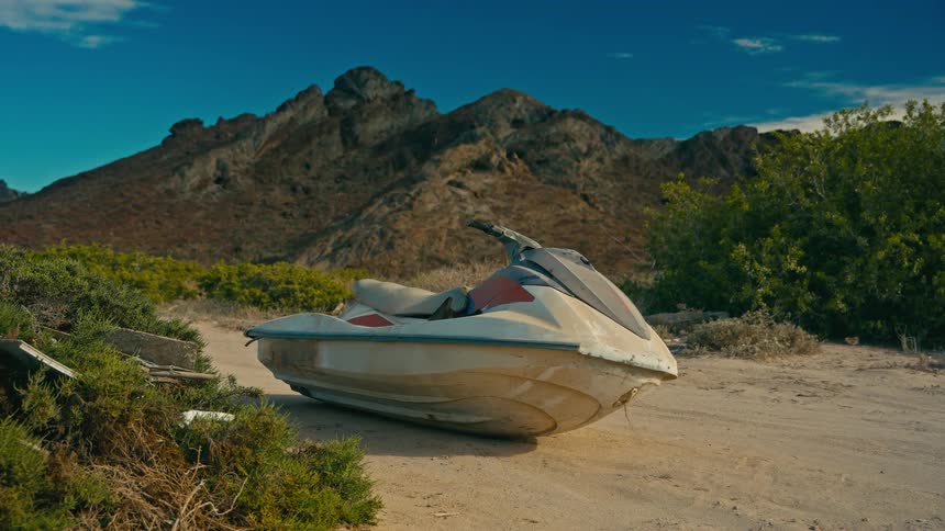 An old, abandoned jet ski rests on a sandy path in a desolate landscape with mountains in the background