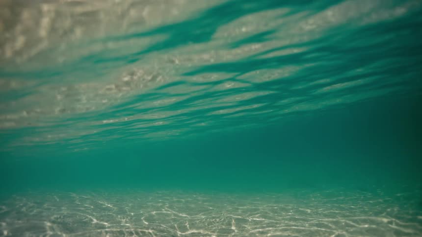 An underwater view of sunlight dancing on the sandy ocean floor, tilting up to the shimmering surface