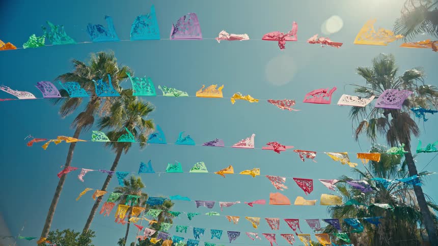 Colorful papel picado banners wave gently in the breeze between palm trees against a bright blue sunny sky