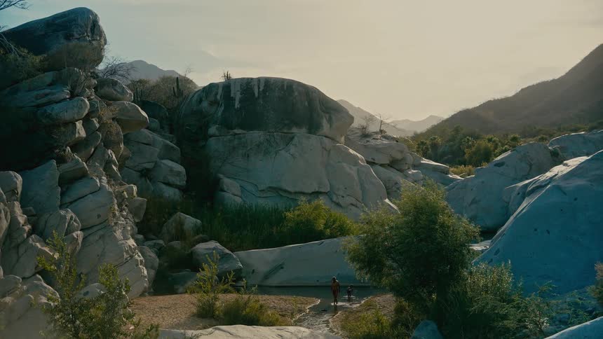 A father and his young son stand together in a tranquil river flowing through a vast, rocky canyon at sunrise