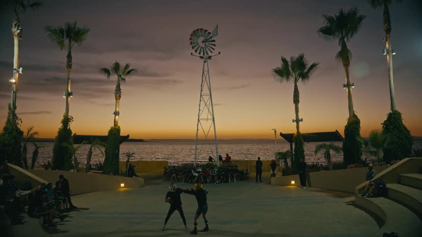 Two fighters spar in a scenic coastal amphitheater with a windmill at sunset as a crowd watches