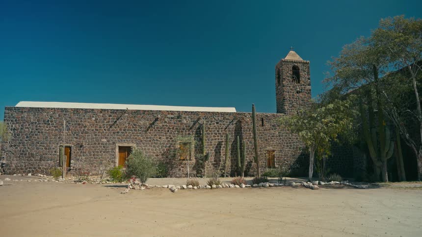 A wide, still shot of an old stone mission surrounded by cacti in a desert landscape under a clear blue sky