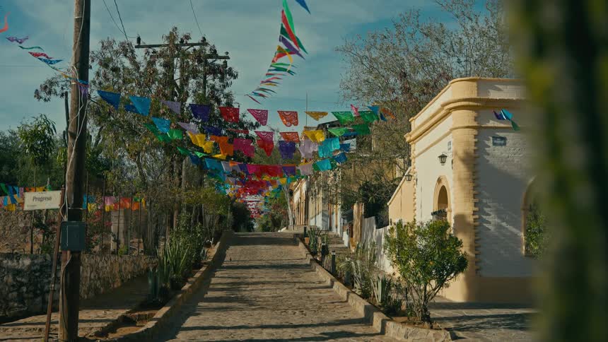 Colorful festive 'papel picado' banners sway gently over a quiet cobblestone street in a traditional Mexican village