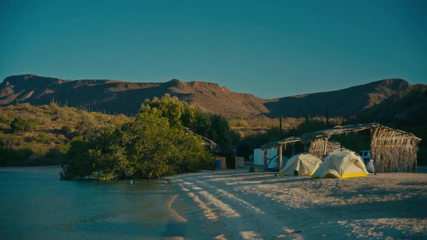 A tranquil campsite with tents is set up on a sandy beach next to a calm bay with desert mountains in the background