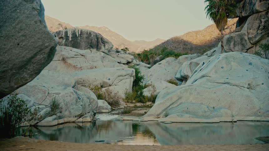 A serene desert oasis with a tranquil pool of water surrounded by large, smooth boulders and distant mountains