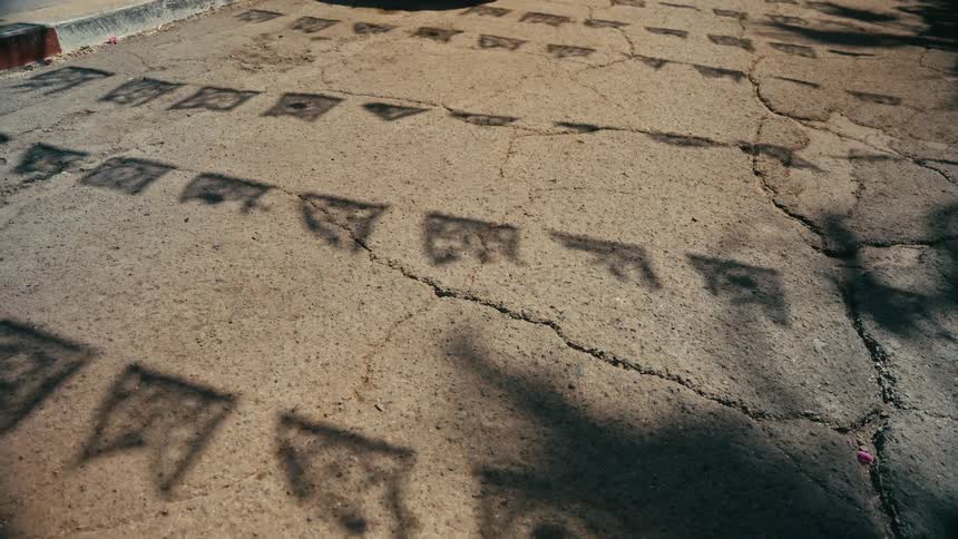 Shadows of swaying Papel Picado banners dance across cracked asphalt on a sunny day