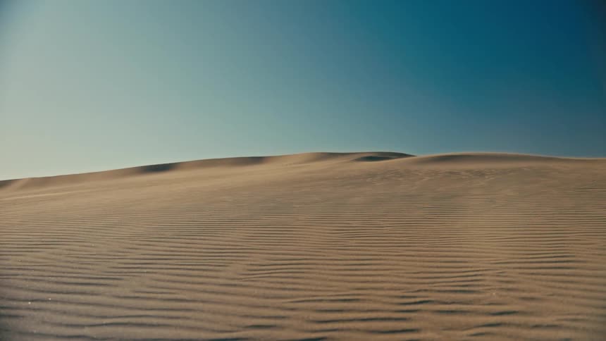 A serene landscape shot of wind blowing over golden sand dunes against a clear blue sky