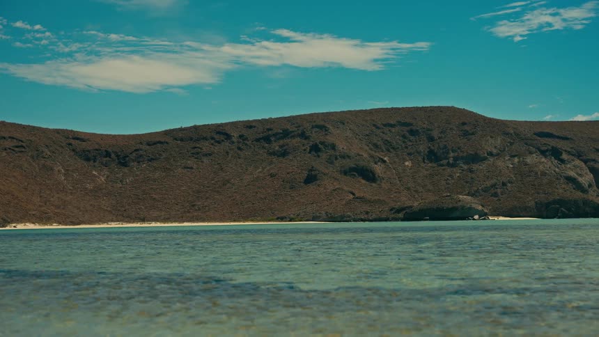 A stunning wide shot captures the tranquil, clear turquoise water of a remote bay with an arid, hilly coastline in the background