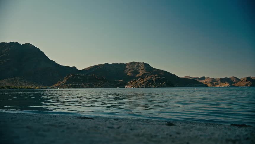 Calm lake waters gently lap the shore with a view of desert mountains under a clear sky