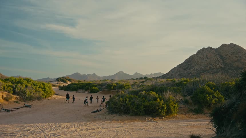 A group of young men play a spirited game of soccer in a wide, sandy clearing surrounded by desert mountains and green brush