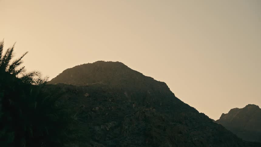 A majestic mountain peak is silhouetted against a hazy golden sky at sunrise or sunset