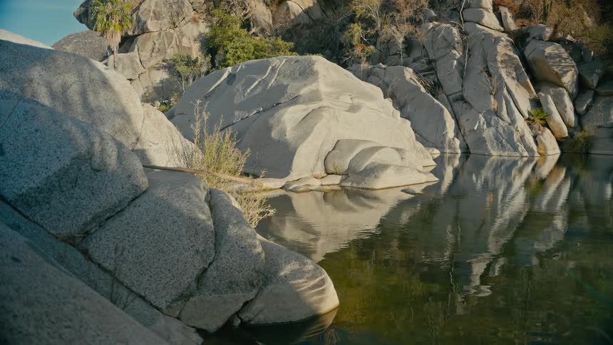 A serene view of a calm, reflecting pool of water surrounded by large, sunlit rock formations in a tranquil canyon