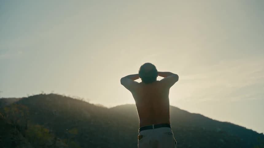 A shirtless man stretches and relaxes as he watches the sun rise over the mountains