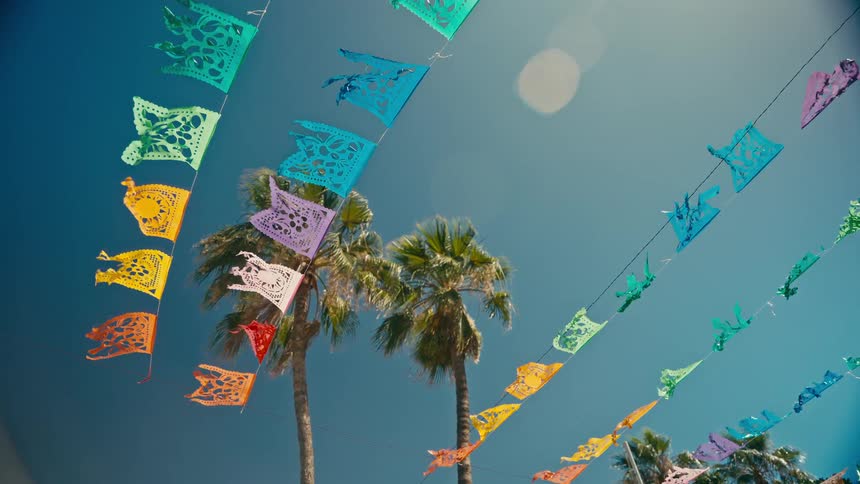 Festive Mexican papel picado banners and tall palm trees wave in the wind against a sunny blue sky