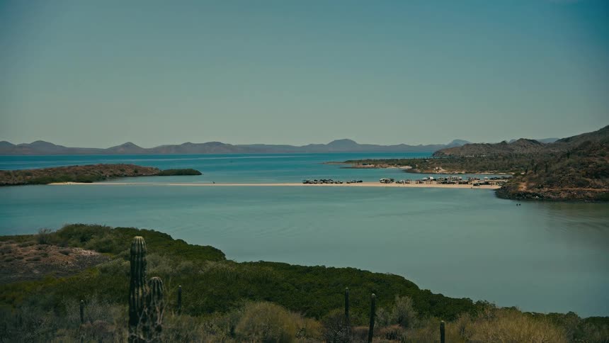A wide shot of a small fishing village on a sandbar in a desert bay, with cacti in the foreground