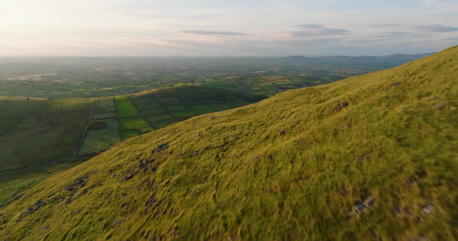 A cinematic aerial shot rises over a grassy, sun-kissed mountainside to reveal a beautiful patchwork of green fields and rolling hills