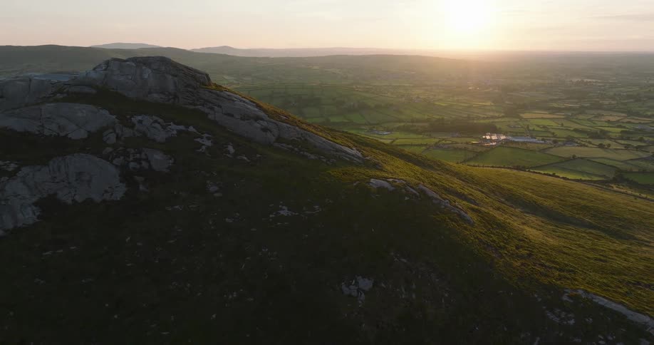 Aerial drone footage flying over a rocky green mountain at sunset, revealing a beautiful patchwork of fields and farmland below
