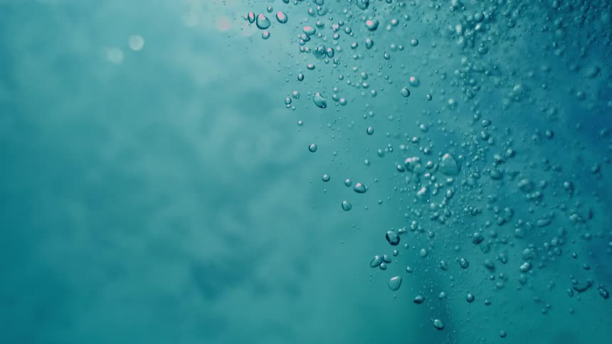 An immersive underwater perspective shows turbulent blue water calming as air bubbles slowly rise towards the sunlit surface