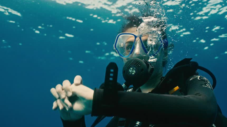 A female scuba diver breathes through her regulator underwater, turning to face the camera in a serene slow-motion shot