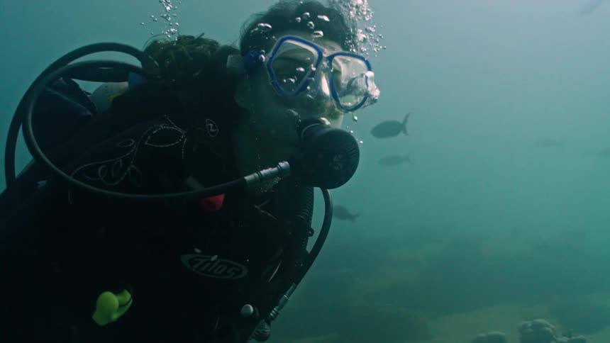 A female scuba diver breathes underwater in slow motion as bubbles rise from her regulator and fish swim in the background