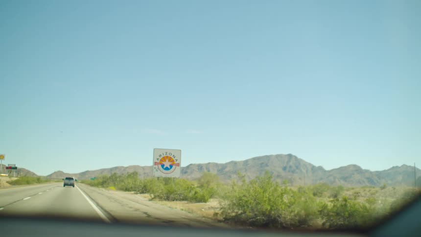 A car drives on a desert highway, passing a 'Welcome to Arizona' sign with mountains in the background
