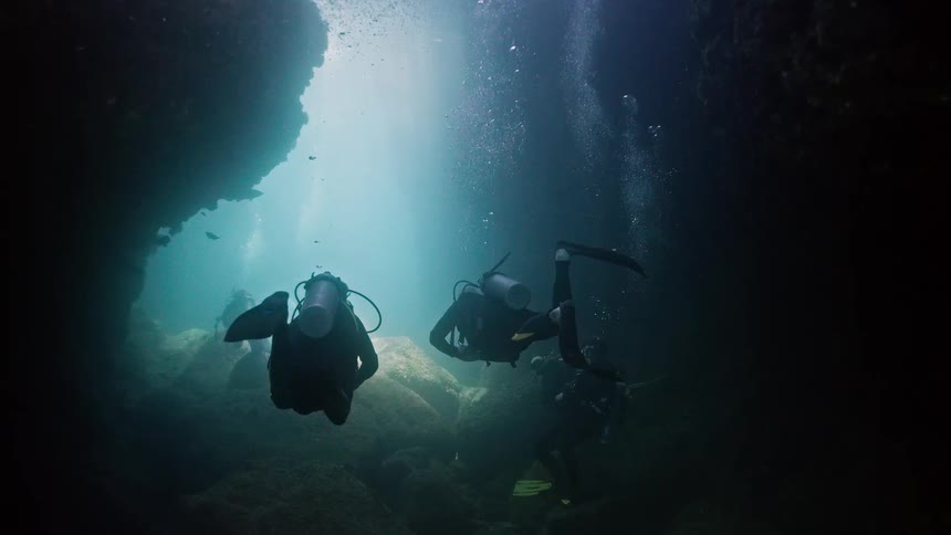A group of scuba divers explore the mysterious depths of an underwater cave, illuminated by a single powerful flashlight