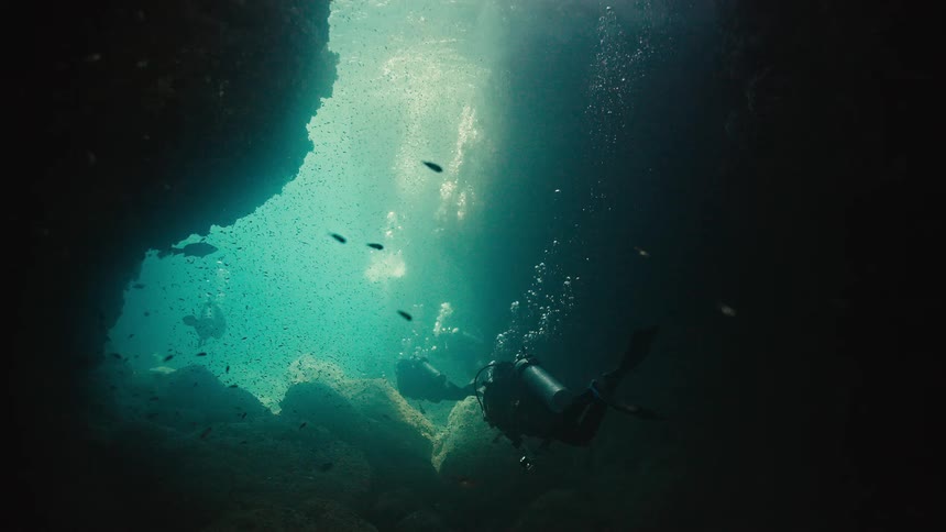 Scuba divers explore a sunlit underwater cave teeming with a school of tiny fish