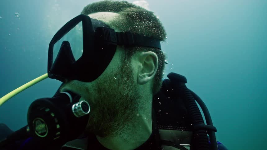 A close-up slow-motion shot of a scuba diver breathing underwater as bubbles rise to the surface